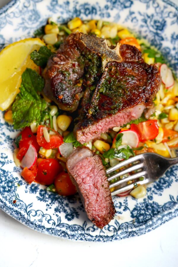 Lamb loin chops grilled in a compound herb butter on top of a tomato salad. There is a piece of the lamb cut into showing this recipe has cooked it to a medium-rare.