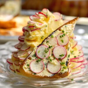 A whipped butter with chives and flaky salt with thin slices of radishes all around sitting on a plate. A piece of toast with the butter spread on it is sitting in front.