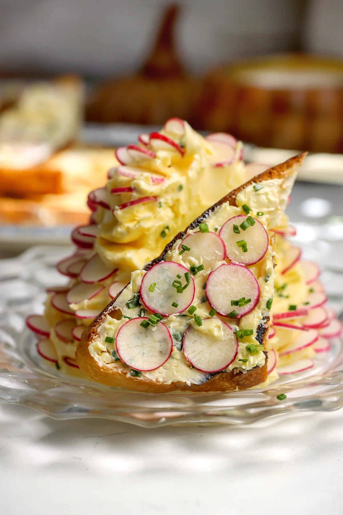 A whipped butter with chives and flaky salt with thin slices of radishes all around sitting on a plate. A piece of toast with the butter spread on it is sitting in front.