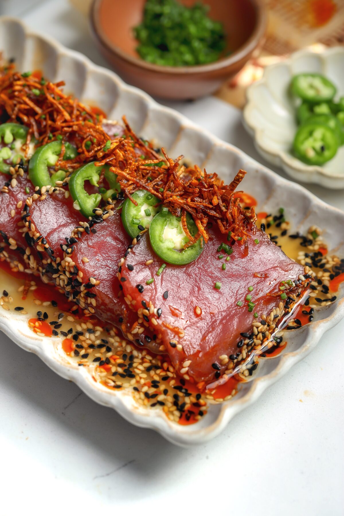 Sesame-crusted tuna crudo with white soy ponzu sauce, topped with crispy carrots and chives on a plate.