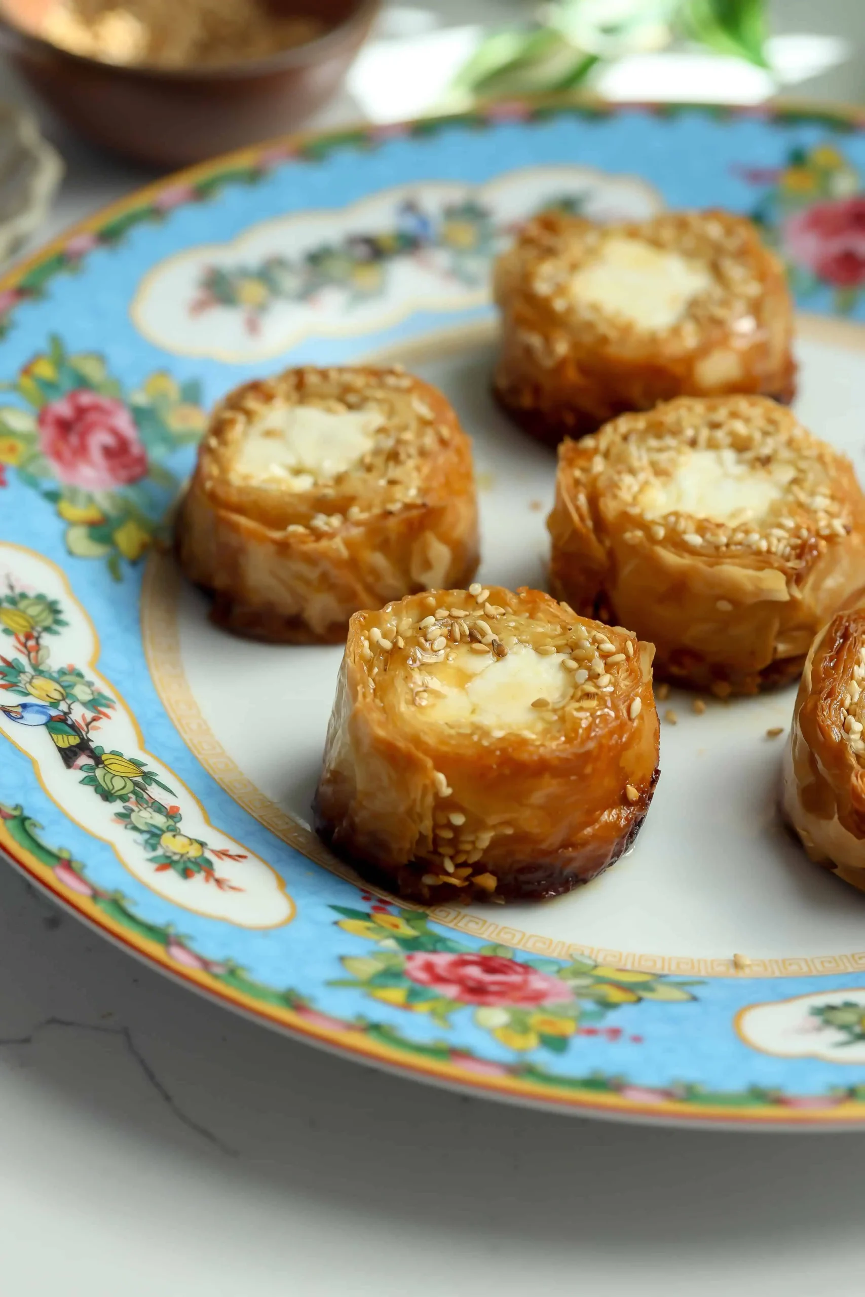 Side angle of Greek-inspired baked feta phyllo bites with honey butter and sesame seeds appetizer on a blue floral plate.