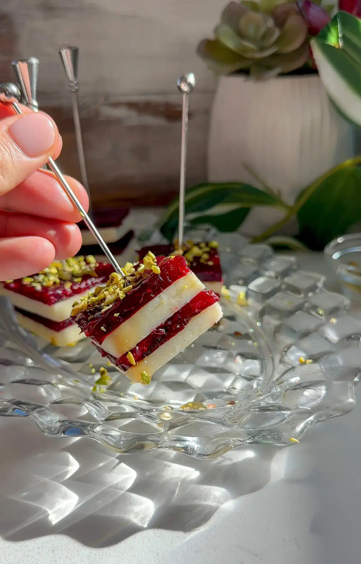Hand holding a stacked cranberry cheddar bite on a silver pick, showing layers of white cheddar and cranberry jelly with chopped pistachios.