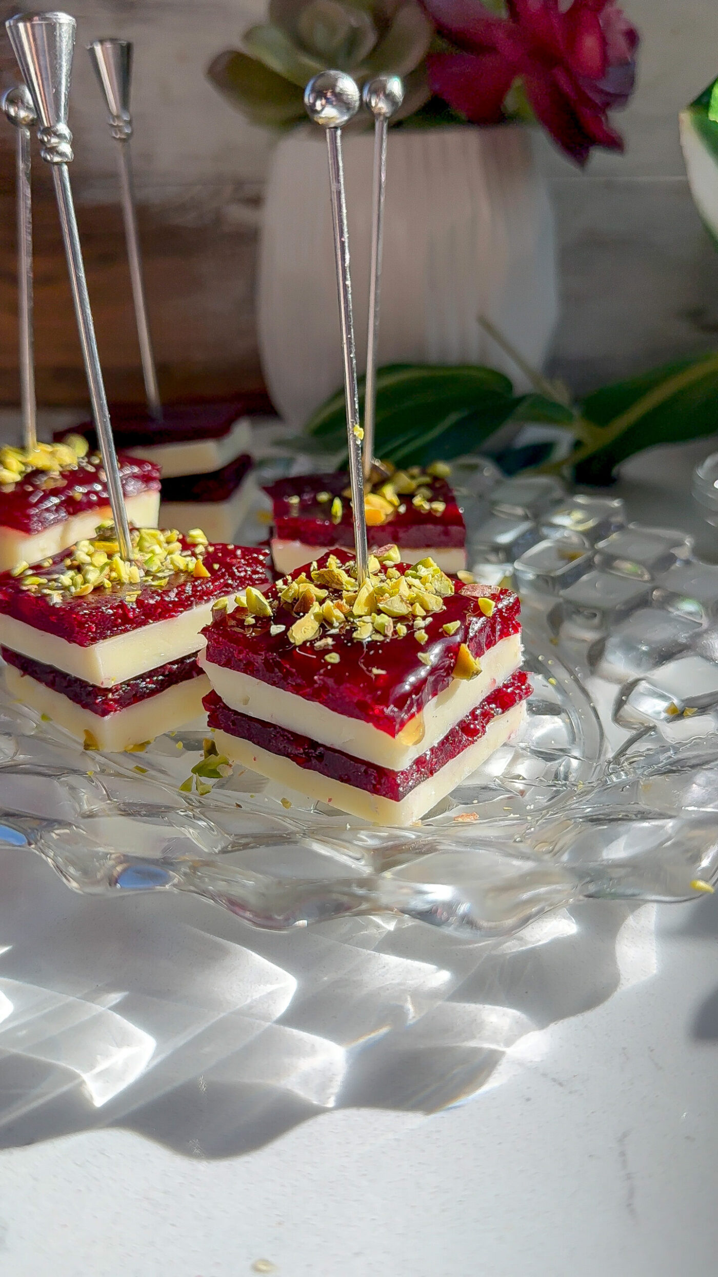 Layered cranberry-cheddar appetizer bites topped with crushed pistachios, served on a clear glass plate with silver picks.