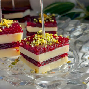 A single layered appetizer bite of sharp white cheddar, cranberry sauce, and cranberry jelly topped with pistachios on a clear glass plate.