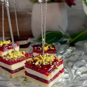 Stacked cranberry cheddar bites with white cheddar and cranberry jelly layers topped with chopped pistachios, on silver picks on a glass plate.