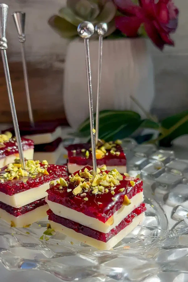 Stacked cranberry cheddar bites with white cheddar and cranberry jelly layers topped with chopped pistachios, on silver picks on a glass plate.