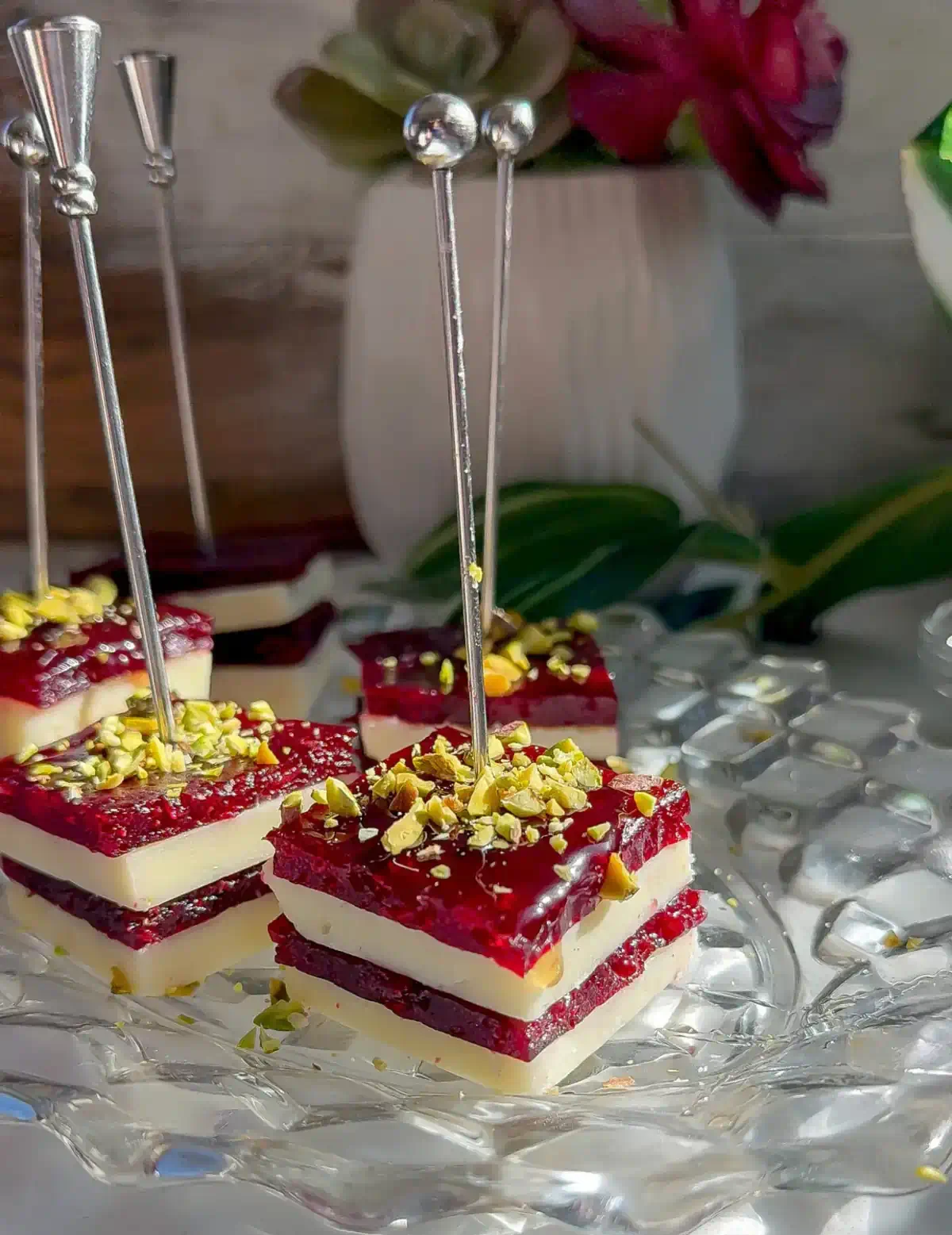 Stacked cranberry cheddar bites with white cheddar and cranberry jelly layers topped with chopped pistachios, on silver picks on a glass plate.
