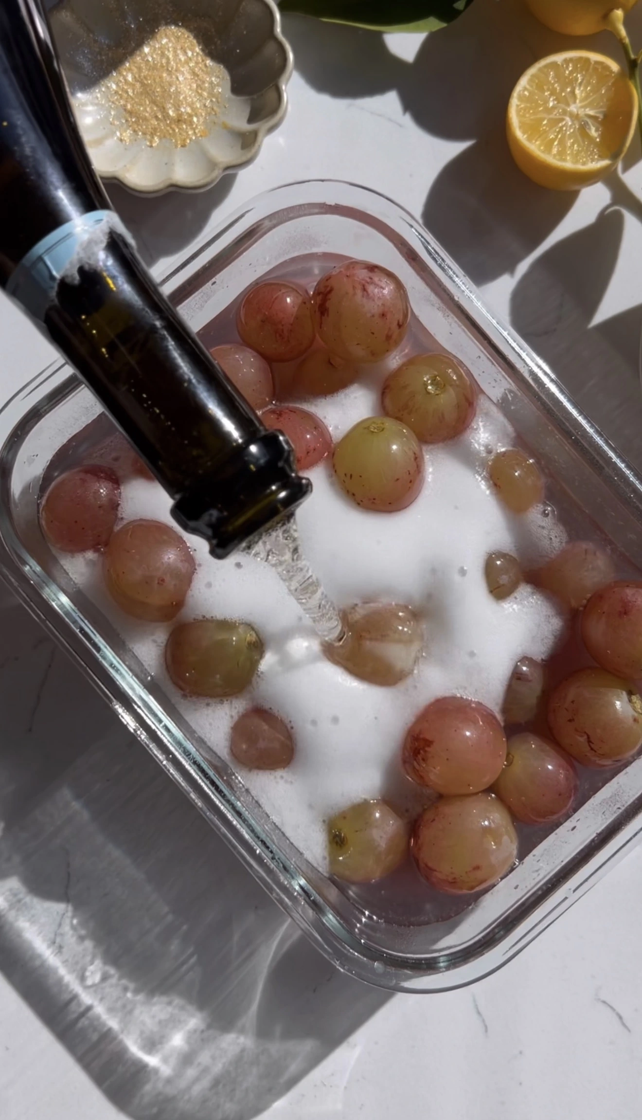 Grapes soaking in champagne and gin in a glass dish with lemon and edible gold nearby.