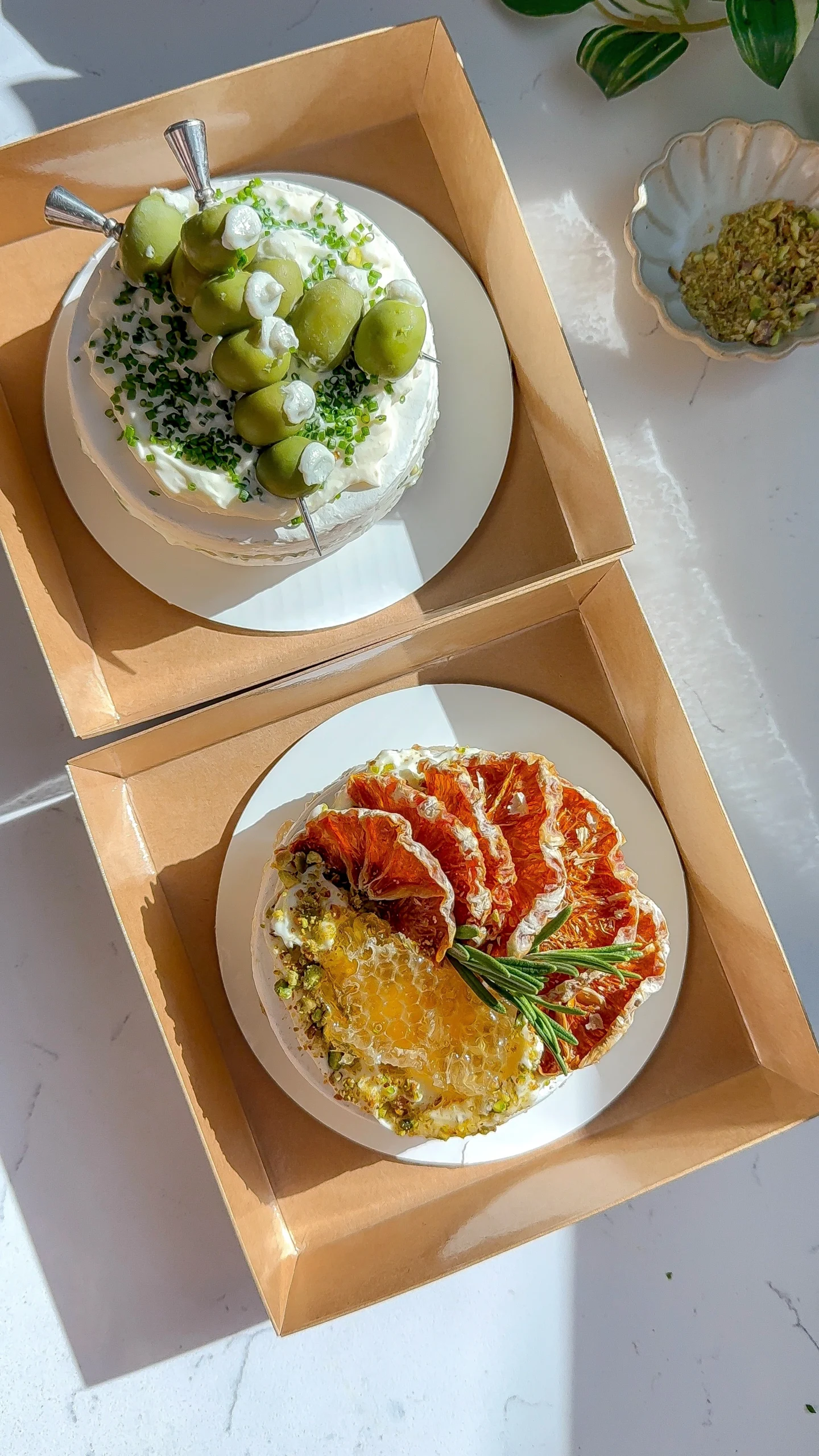 Two brie cakes presented in gift boxes, one topped with blue cheese stuffed olives and chives and the other with citrus, honeycomb, and pistachios.