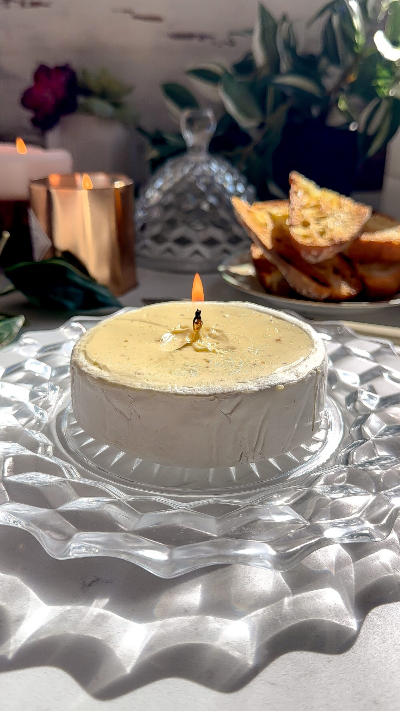 A roasted garlic brie butter candle with a lit food-safe wick, set on a glass plate with toasted bread in the background.