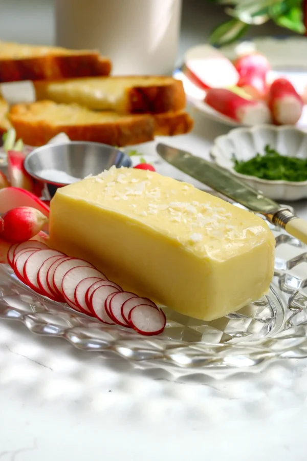 Cultured butter served with sliced radishes and flaky sea salt on a glass platter with bread in the background.
