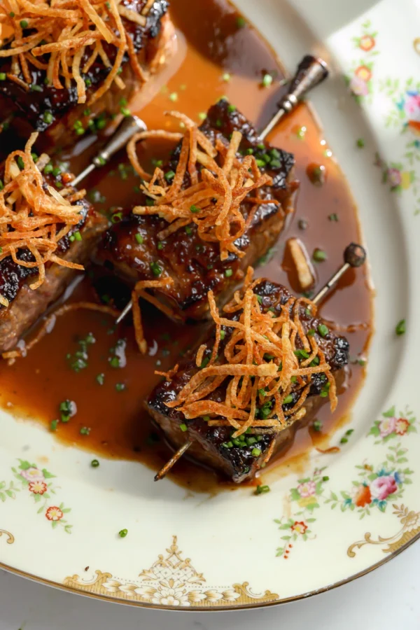 Top-down view of four ribeye appetizer skewers glazed in a savory miso reduction and topped with golden-brown matchstick fries.