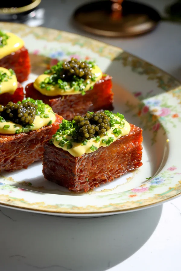 A close-up shot of golden brown crispy potato bricks topped with yellow egg yolk sauce, chives, and black caviar on a floral plate.