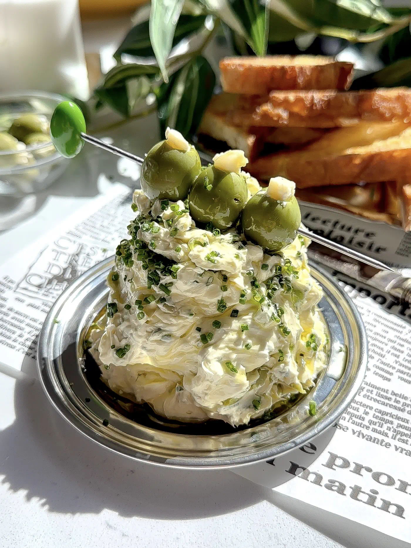 A tall mound of fluffy whipped butter with herbs and blue cheese, garnished with three castelvetrano olives on a skewer, served on a silver plate with toasted bread in the background.