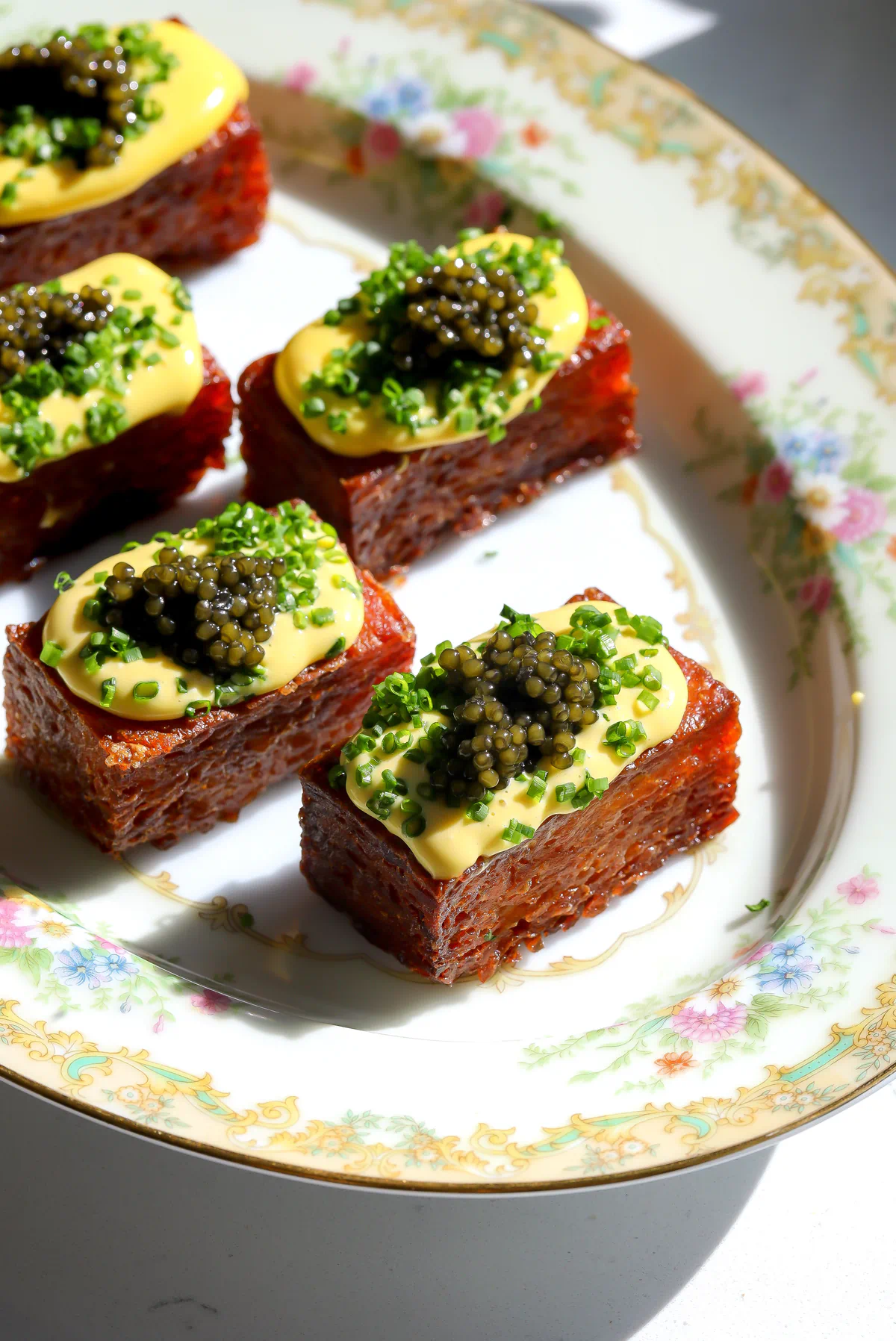 Multiple crispy potato rosti bricks arranged on a vintage floral platter garnished with yellow sauce and caviar.