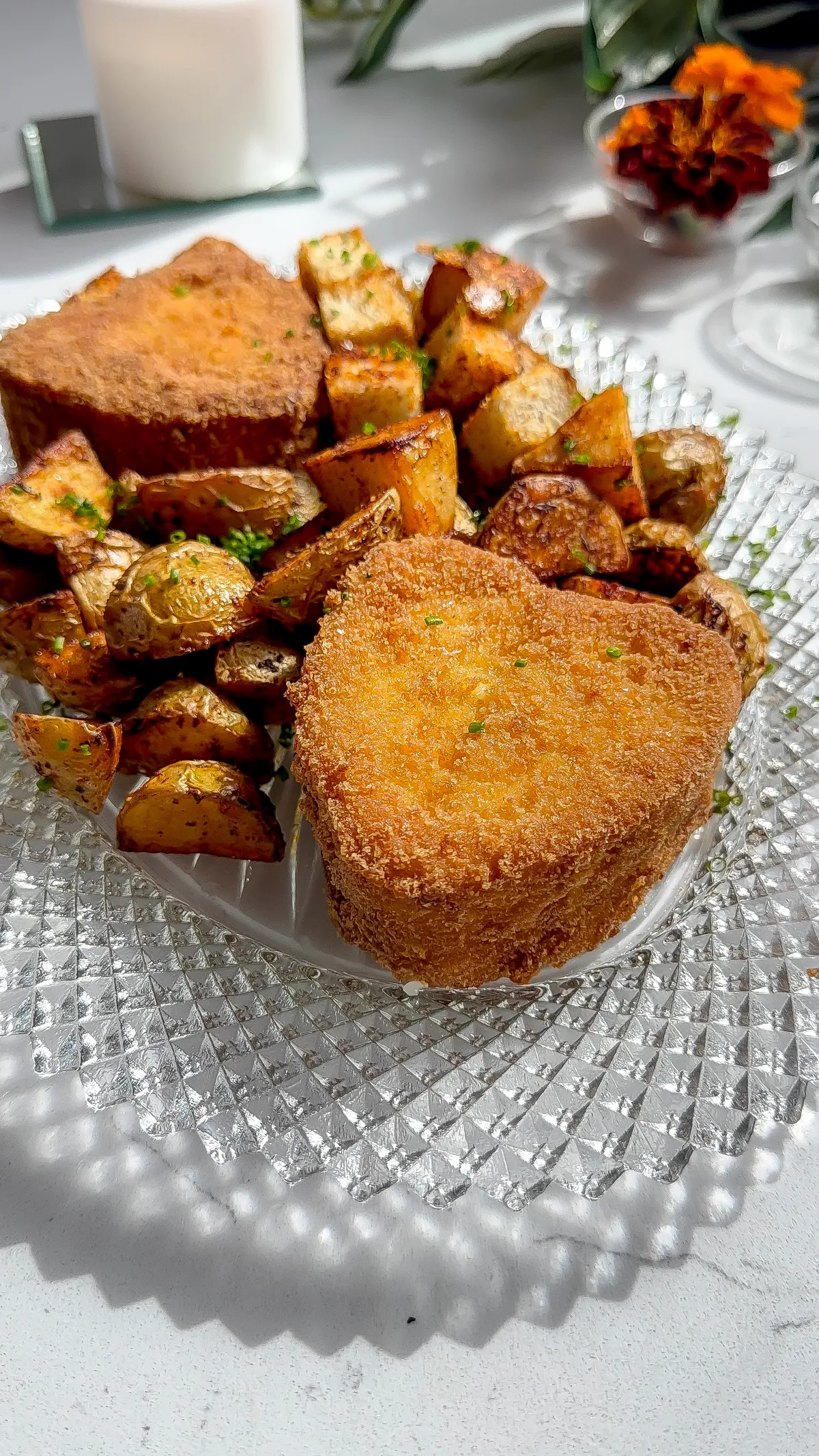 A golden, heart-shaped fried cheese wheel served on a crystal plate alongside roasted peewee potatoes.