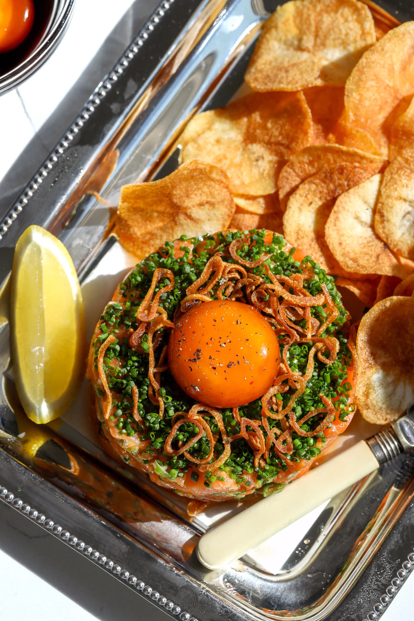 An overhead view of a spicy salmon tartar appetizer on a silver tray, showing the cured egg yolk and crispy toppings with lemon wedges.