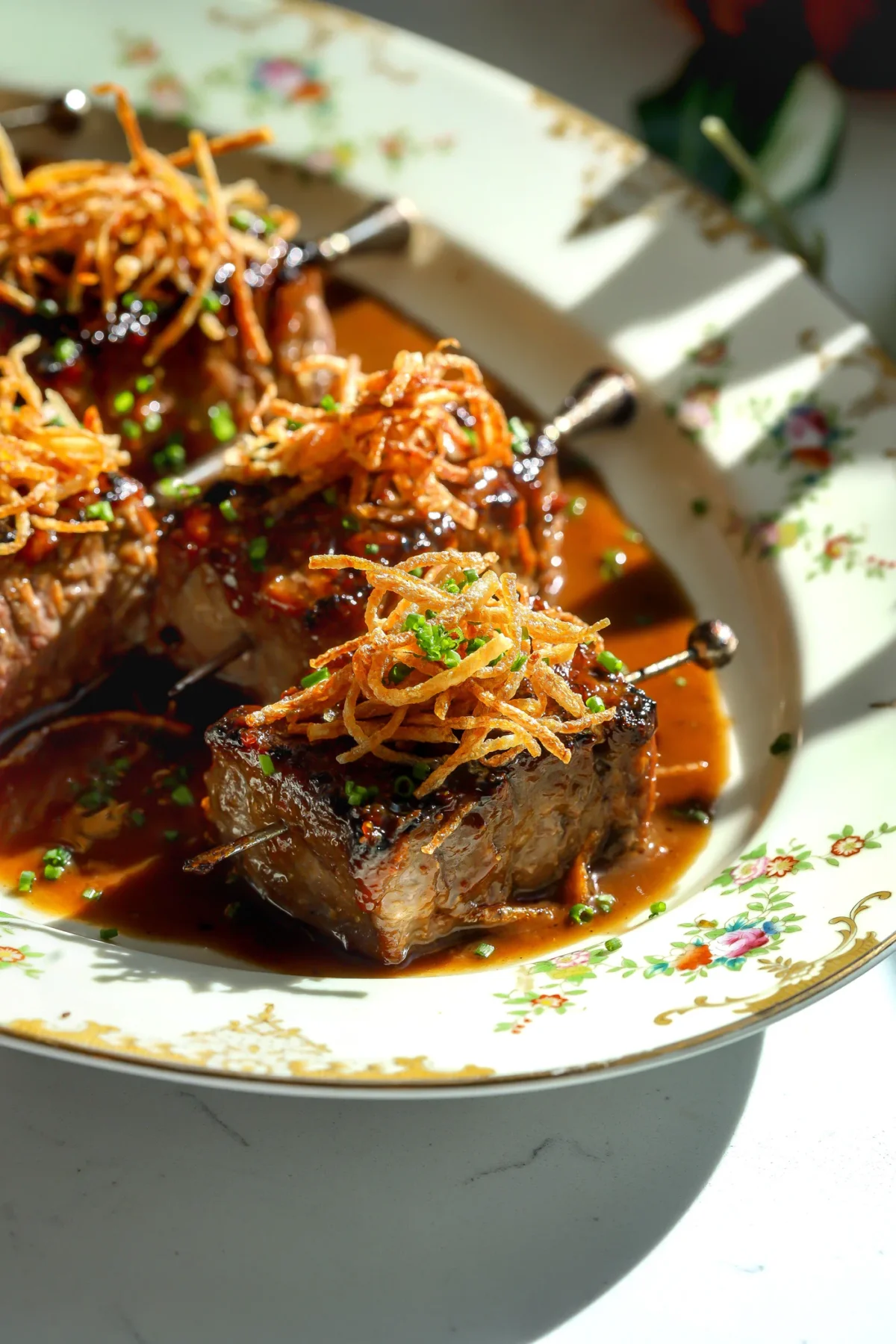 Close-up of layered ribeye steak squares served as appetizers on skewers, topped with crispy potato frites and fresh chives on a vintage platter.