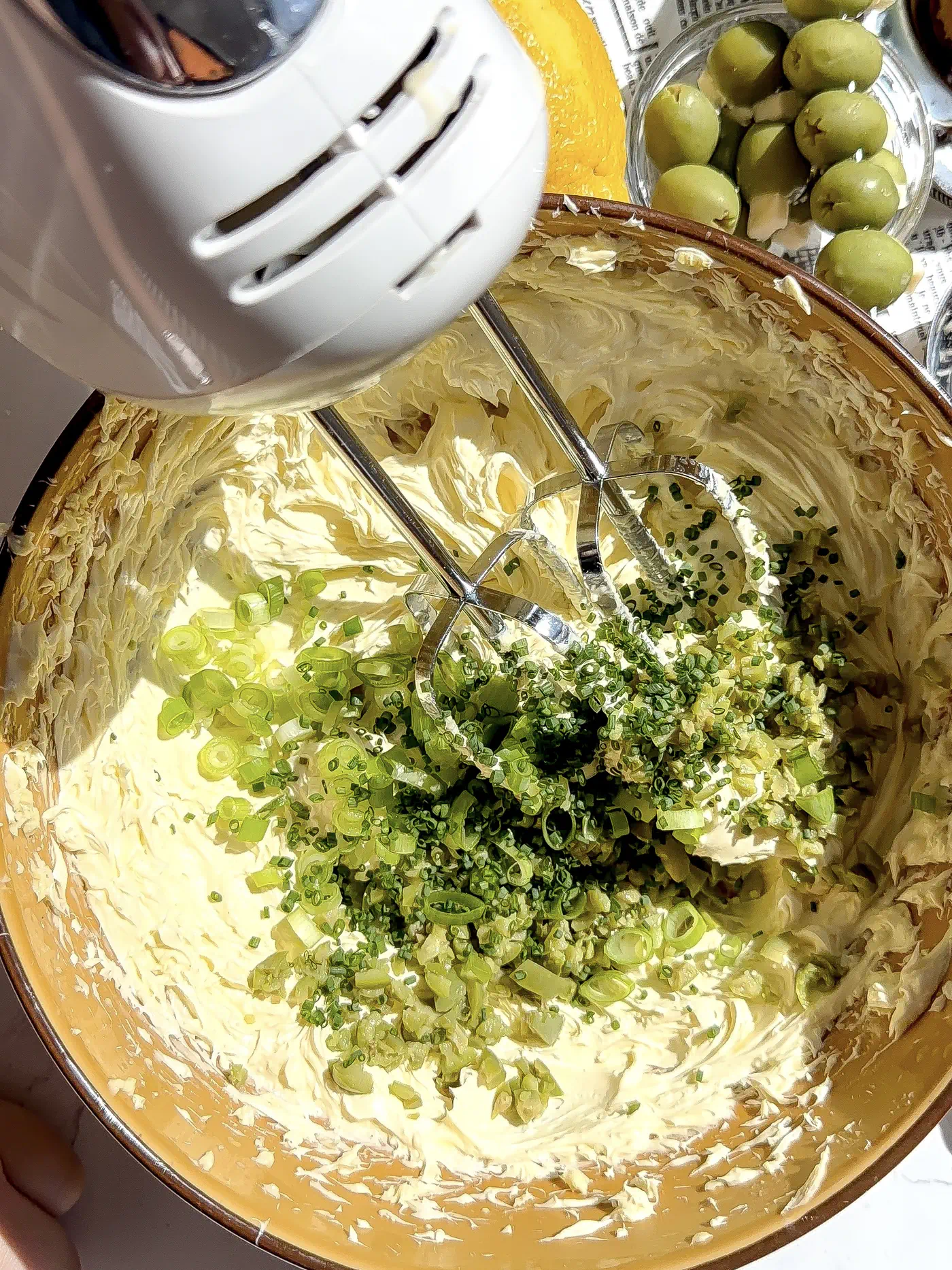 An overhead shot of a hand mixer with whisks folding chopped scallions and herbs into a bowl of pale yellow whipped butter.