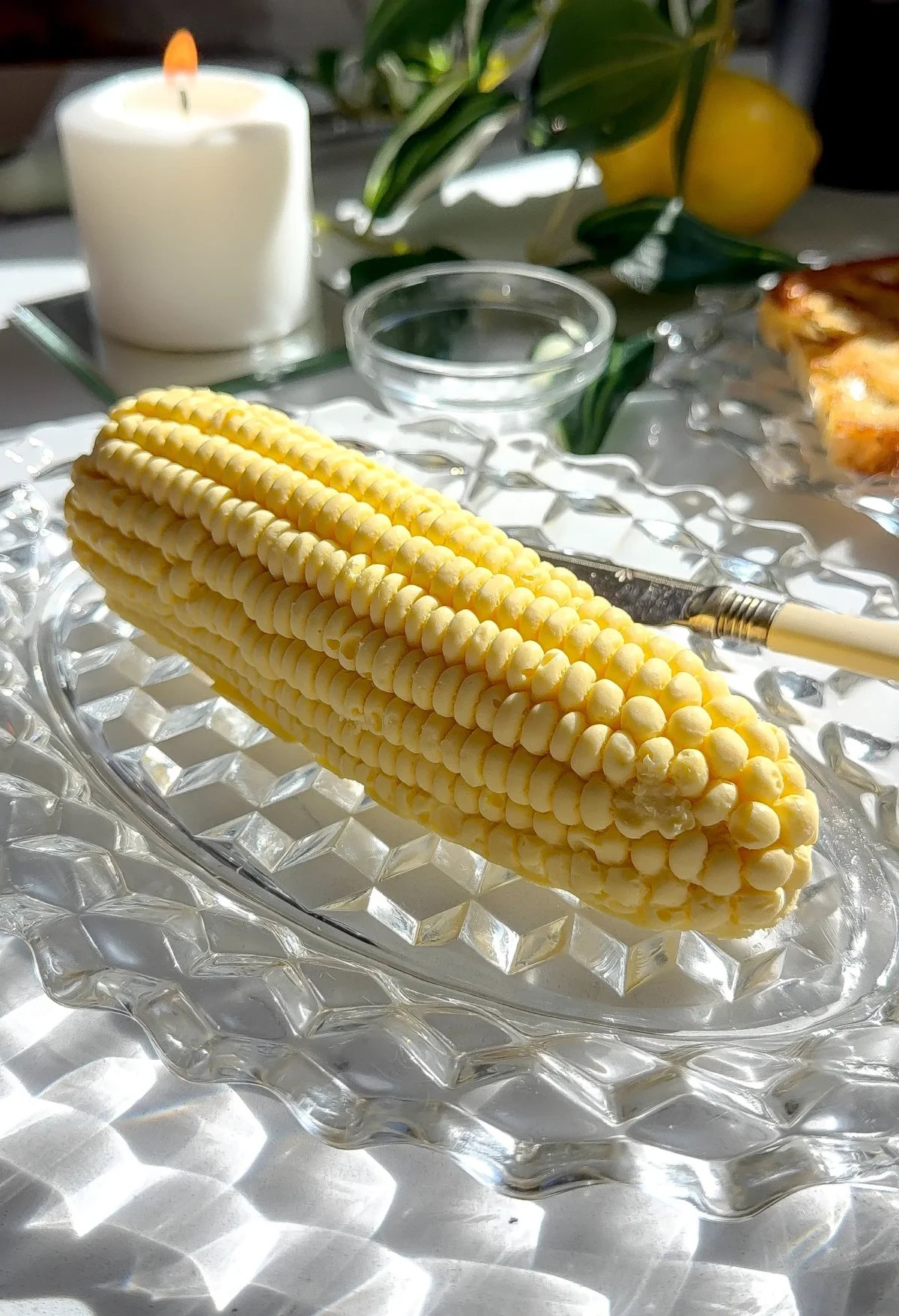 A hyper-realistic ear of corn made from molded compound butter resting on a crystal dish with a lit candle in the background.