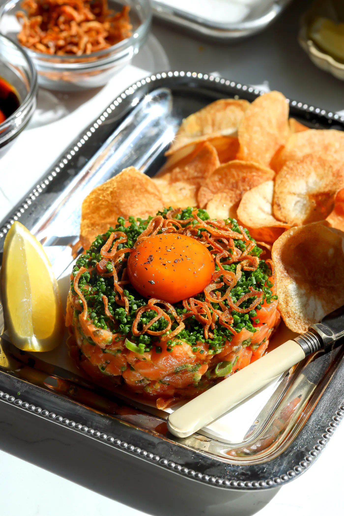 A beautifully plated spicy salmon tartare on a silver platter, garnished with a glossy soy-cured egg yolk, fresh chives, and crispy fried shallots, served with homemade potato chips.