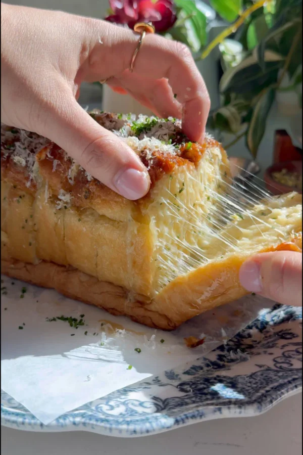 A hand pulling a slice of cheesy garlic pull-apart bread from the loaf, showing a long string of melted sharp white cheddar with a bowl of soup in the background.