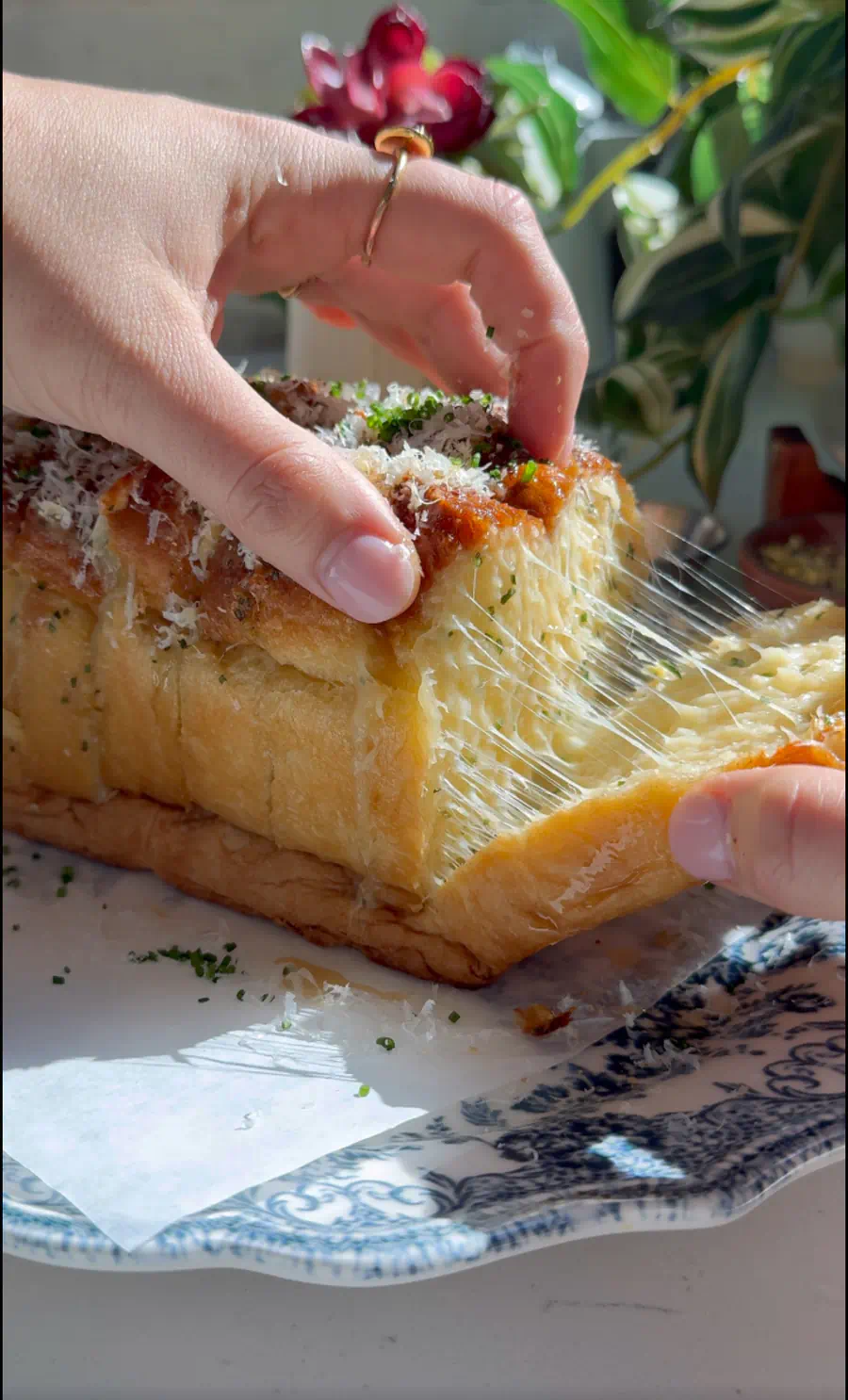 A hand pulling a slice of cheesy garlic pull-apart bread from the loaf, showing a long string of melted sharp white cheddar with a bowl of soup in the background.