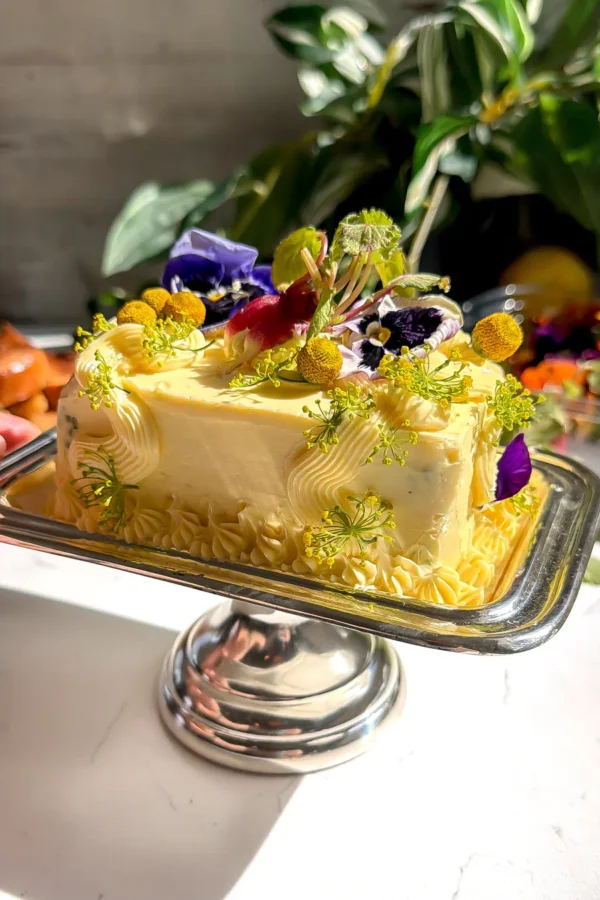 A rectangular herb butter cake on a silver pedestal stand decorated with radishes, purple pansies, and yellow dill flowers.