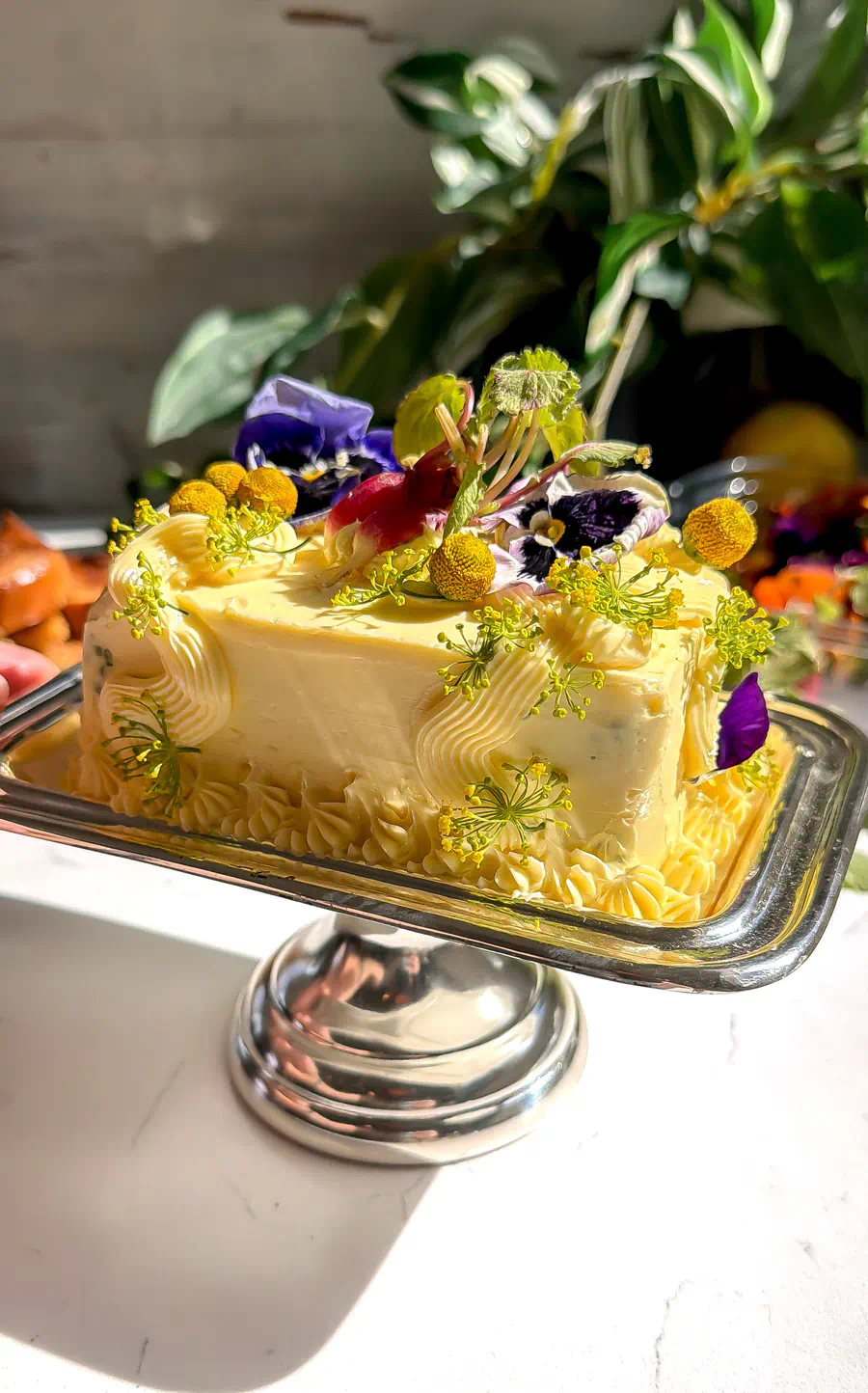 A rectangular herb butter cake on a silver pedestal stand decorated with radishes, purple pansies, and yellow dill flowers.