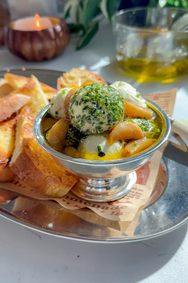 A silver bowl of herbed goat cheese balls with jammy garlic confit cloves and toasted bread on a marble surface.
