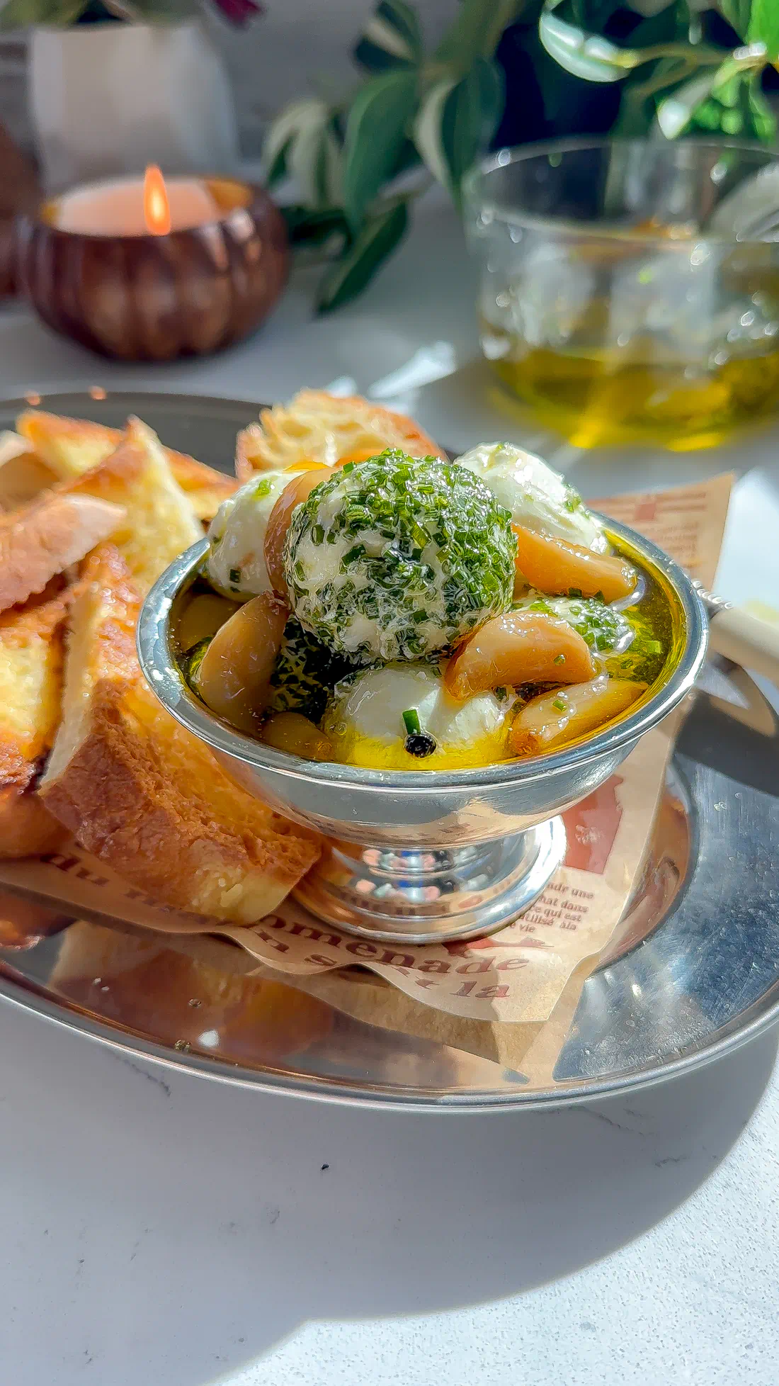 A silver bowl of herbed goat cheese balls with jammy garlic confit cloves and toasted bread on a marble surface.