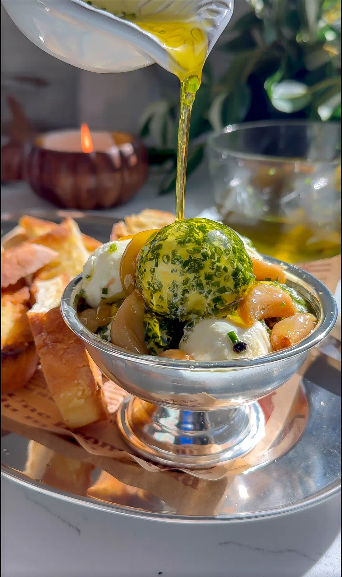 Golden garlic-infused olive oil pouring from a white pitcher onto chilled goat cheese balls coated in fresh chives.