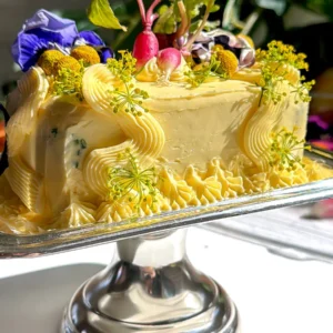Side view of a decorated savory butter cake on a silver stand with a background of green foliage.