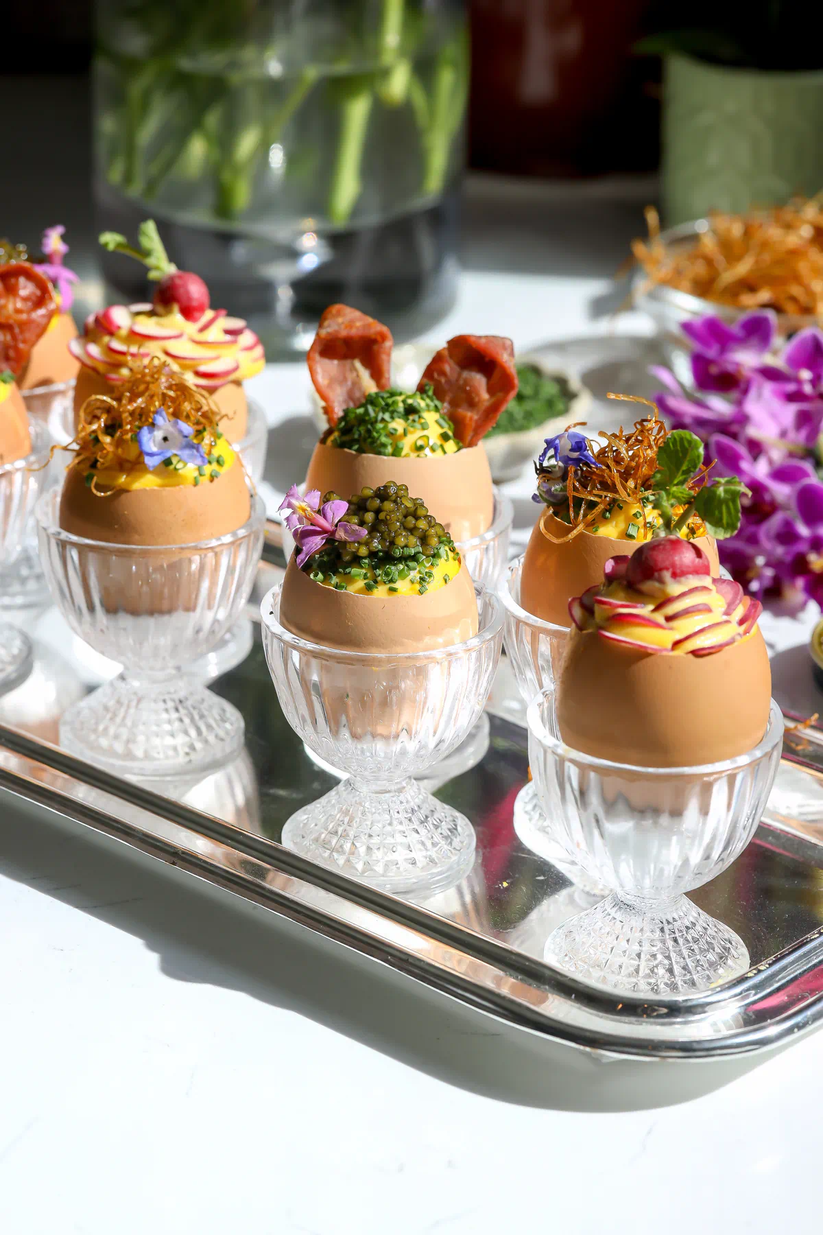 Deviled egg salad inside Egg shells that have been precision cut with an egg topper sitting in glass egg cups. Topped with a 4 flight of topping options.