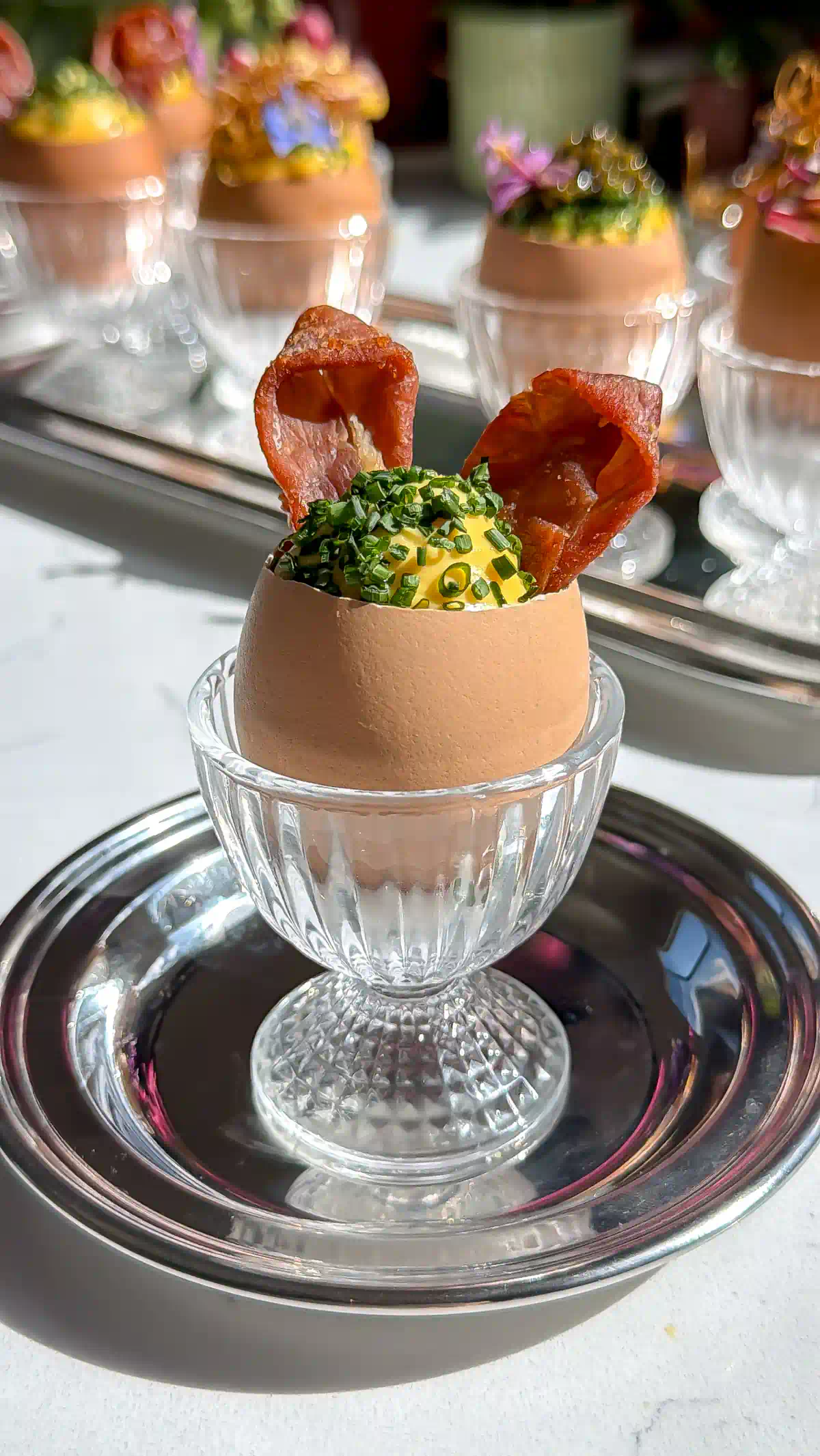 Deviled egg salad inside Egg shells that have been precision cut with an egg topper sitting in glass egg cups. Topped with chives and prosciutto rabbit ears for easter.