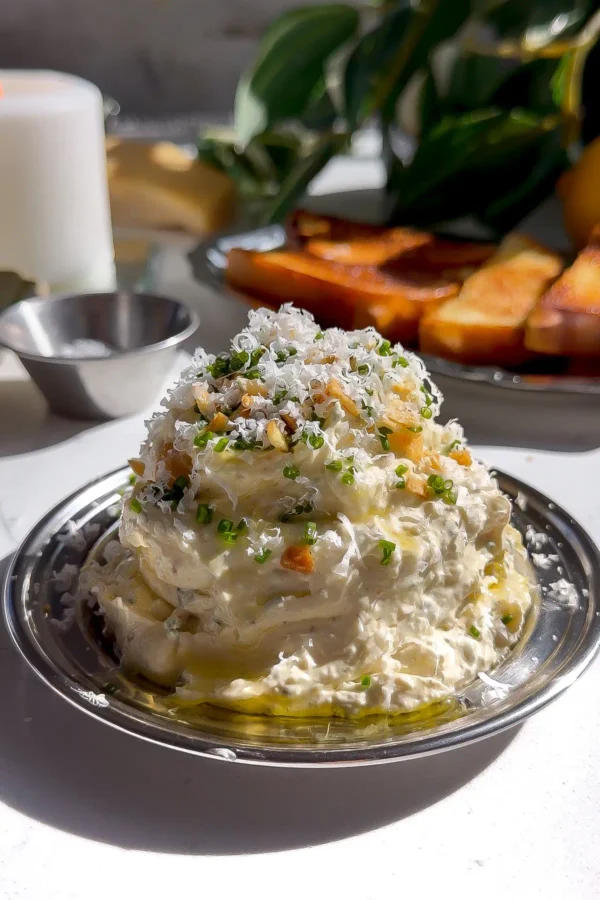 A tall mound of fluffy roasted garlic whipped butter garnished with freshly grated Parmigiano-Reggiano, chopped chives, and crispy fried garlic on a small silver plate.