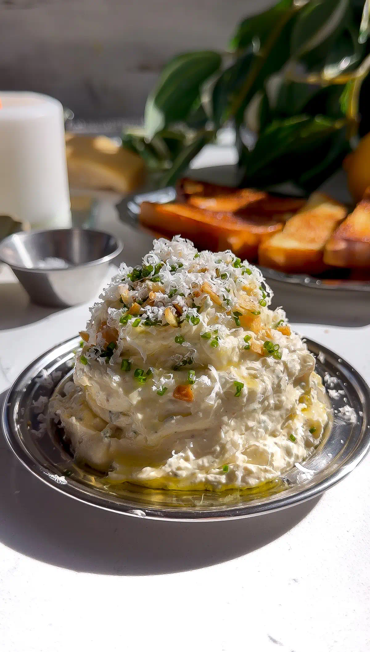 A tall mound of fluffy roasted garlic whipped butter garnished with freshly grated Parmigiano-Reggiano, chopped chives, and crispy fried garlic on a small silver plate.