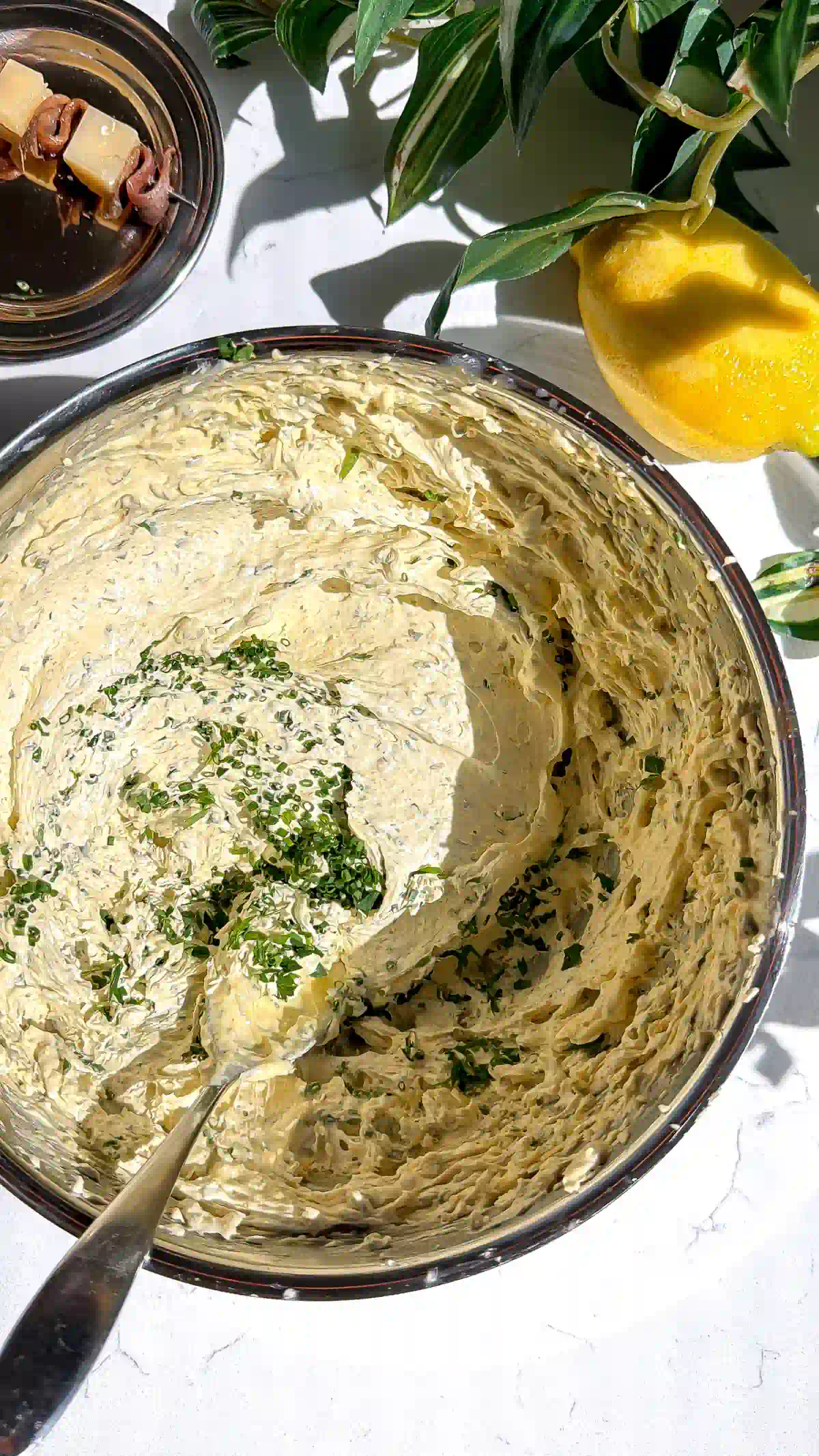 A top-down view of fluffy Caesar whipped brown butter in a metal mixing bowl with a spoon resting inside, sprinkled with fresh chopped chives.