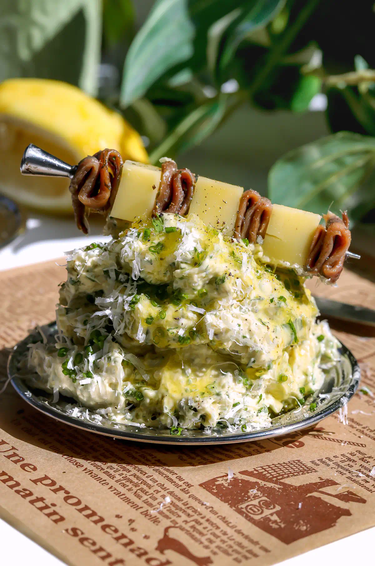 A close-up of Caesar whipped butter piled on a plate over French-script parchment paper, topped with fresh herbs, grated cheese, and an anchovy and parmesan skewer with a lemon in the background.