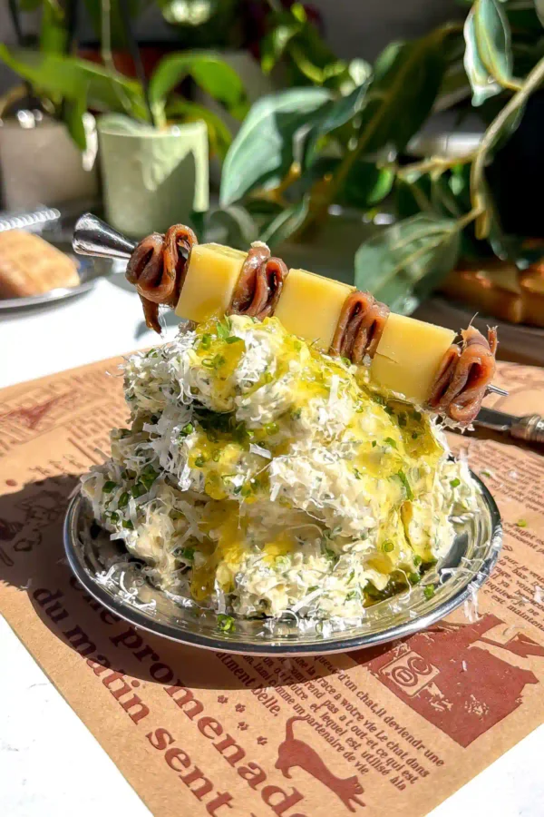Caesar whipped butter piled on a plate over French-script parchment paper, topped with fresh herbs, grated cheese, and an anchovy and parmesan skewer with a lemon in the background.