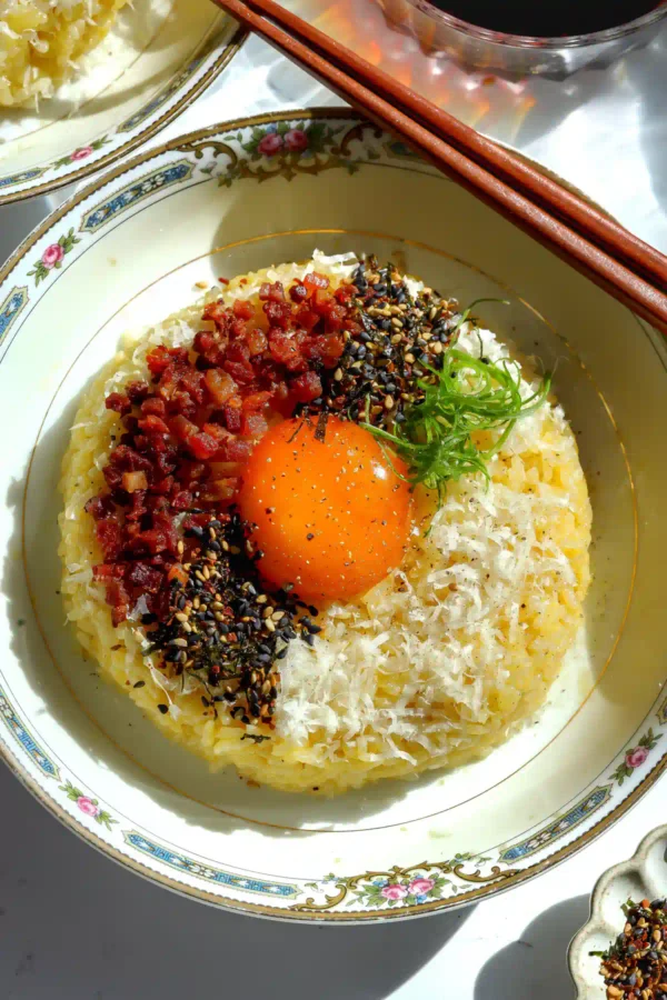 Overhead view of a vibrant bowl of Wafu Carbonara Tamago Kake Gohan (TKG) featuring a glossy raw egg yolk, crispy rendered pancetta, and a snowfall of finely microplaned pecorino cheese.