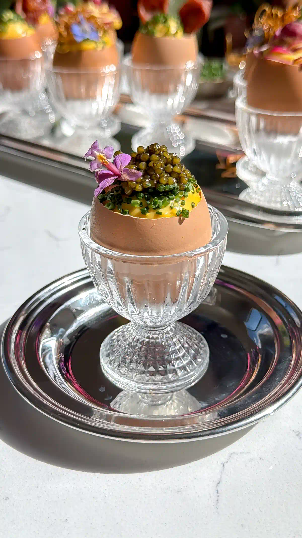 Deviled egg salad inside Egg shells that have been precision cut with an egg topper sitting in glass egg cups. Topped with chives, caviar and an edible flower.