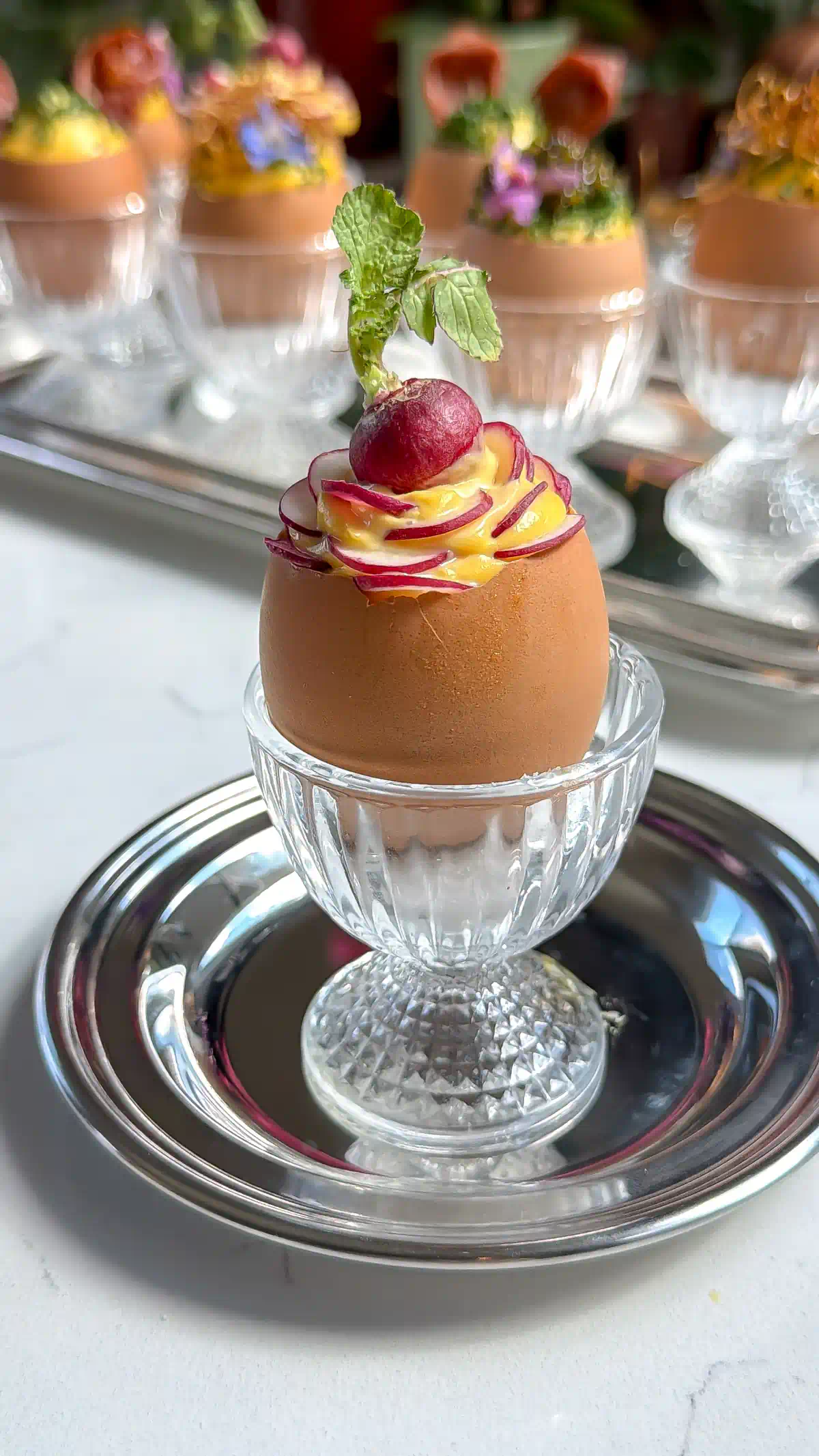 Deviled egg salad inside Egg shells that have been precision cut with an egg topper sitting in glass egg cups. Topped with sliced mini radishes.