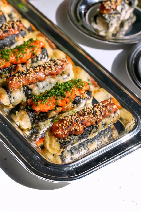 A close-up of crispy tempura sushi taquitos on a silver serving tray, featuring a creamy spicy tuna topping garnished with sesame seeds and a cured spicy salmon topping with fresh chives.