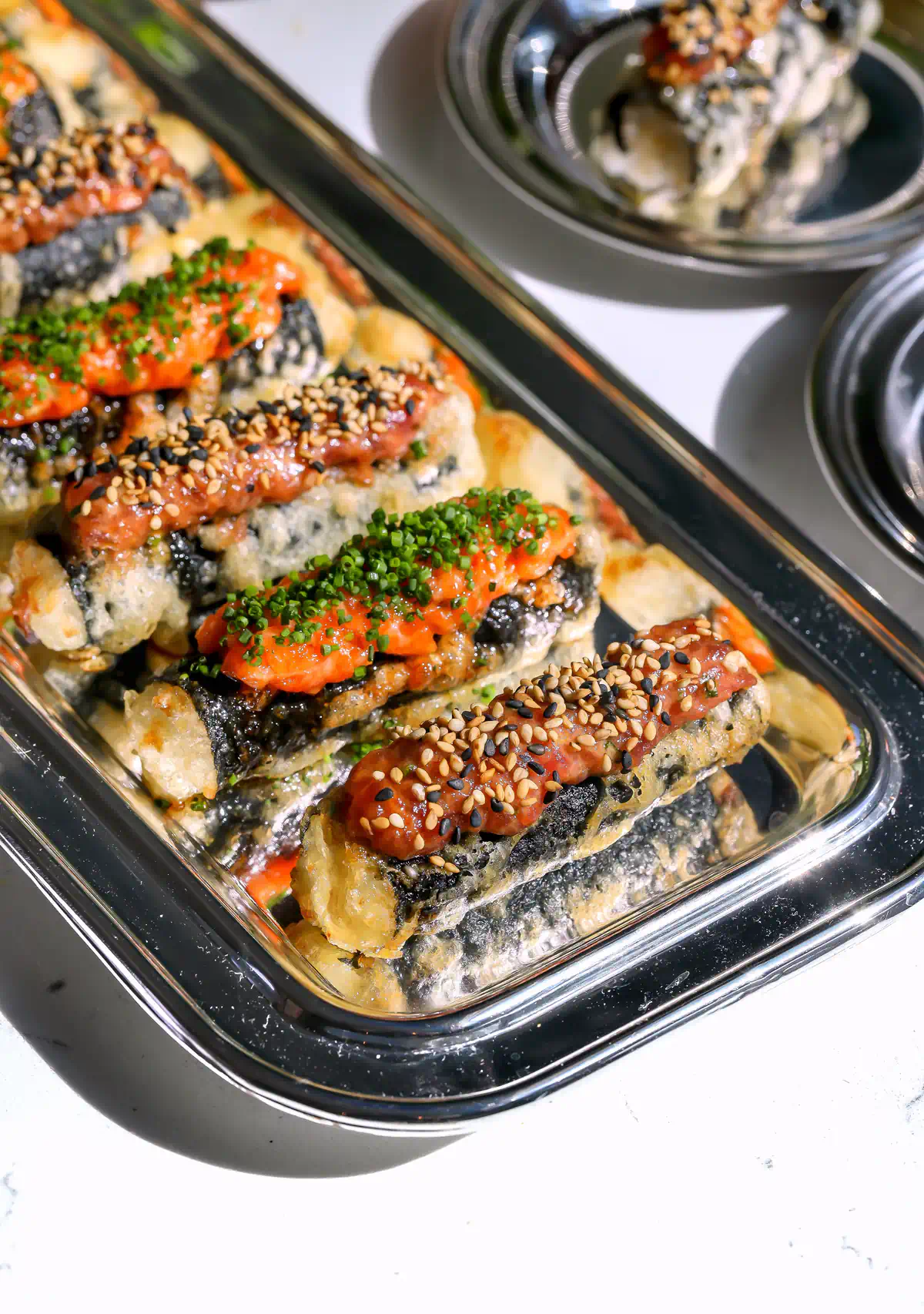 A close-up of crispy tempura sushi taquitos on a silver serving tray, featuring a creamy spicy tuna topping garnished with sesame seeds and a cured spicy salmon topping with fresh chives.
