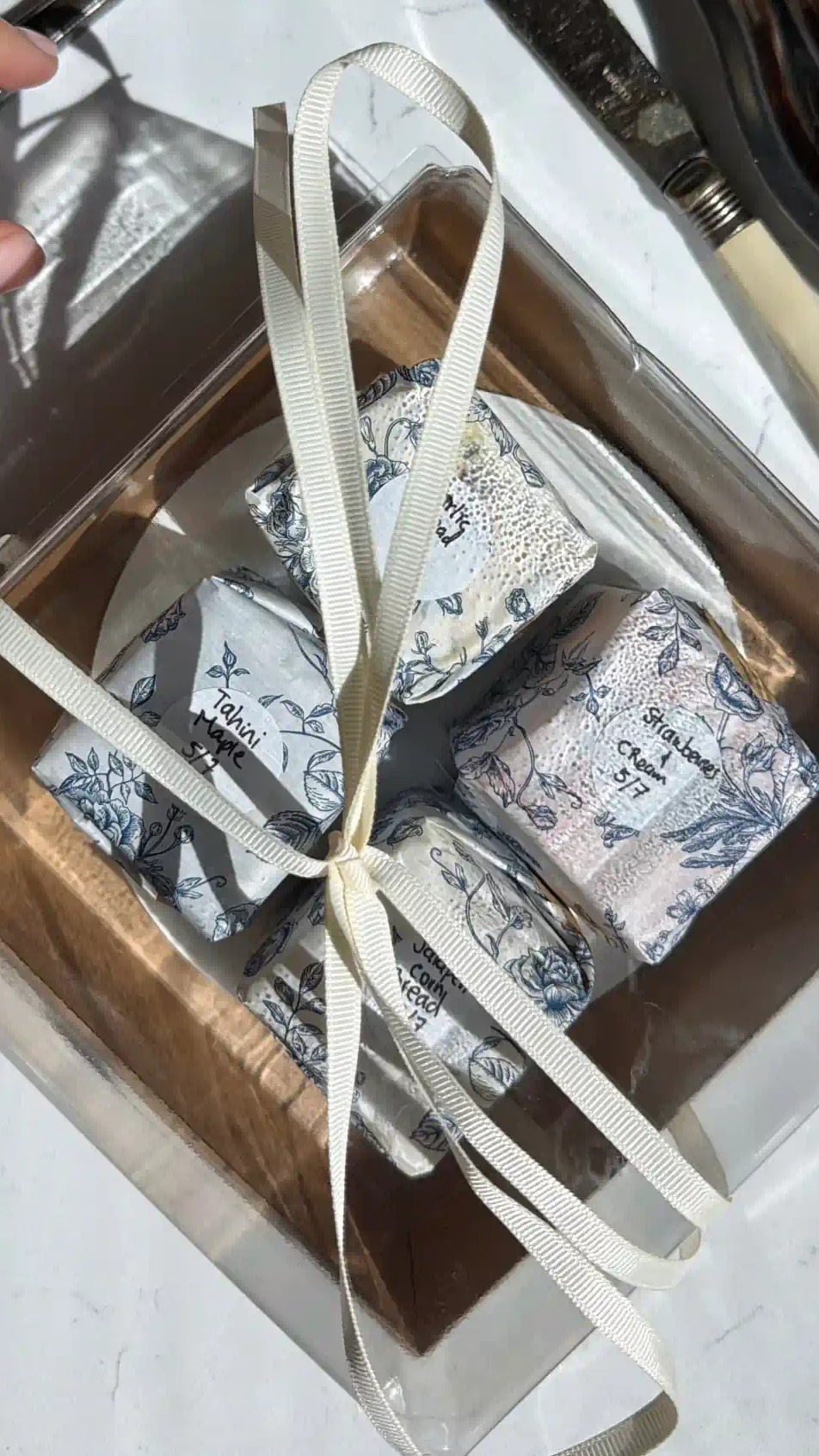 A compound butter flight packaged as a gift, featuring four blocks wrapped in blue floral paper with handwritten labels and tied with a white ribbon.