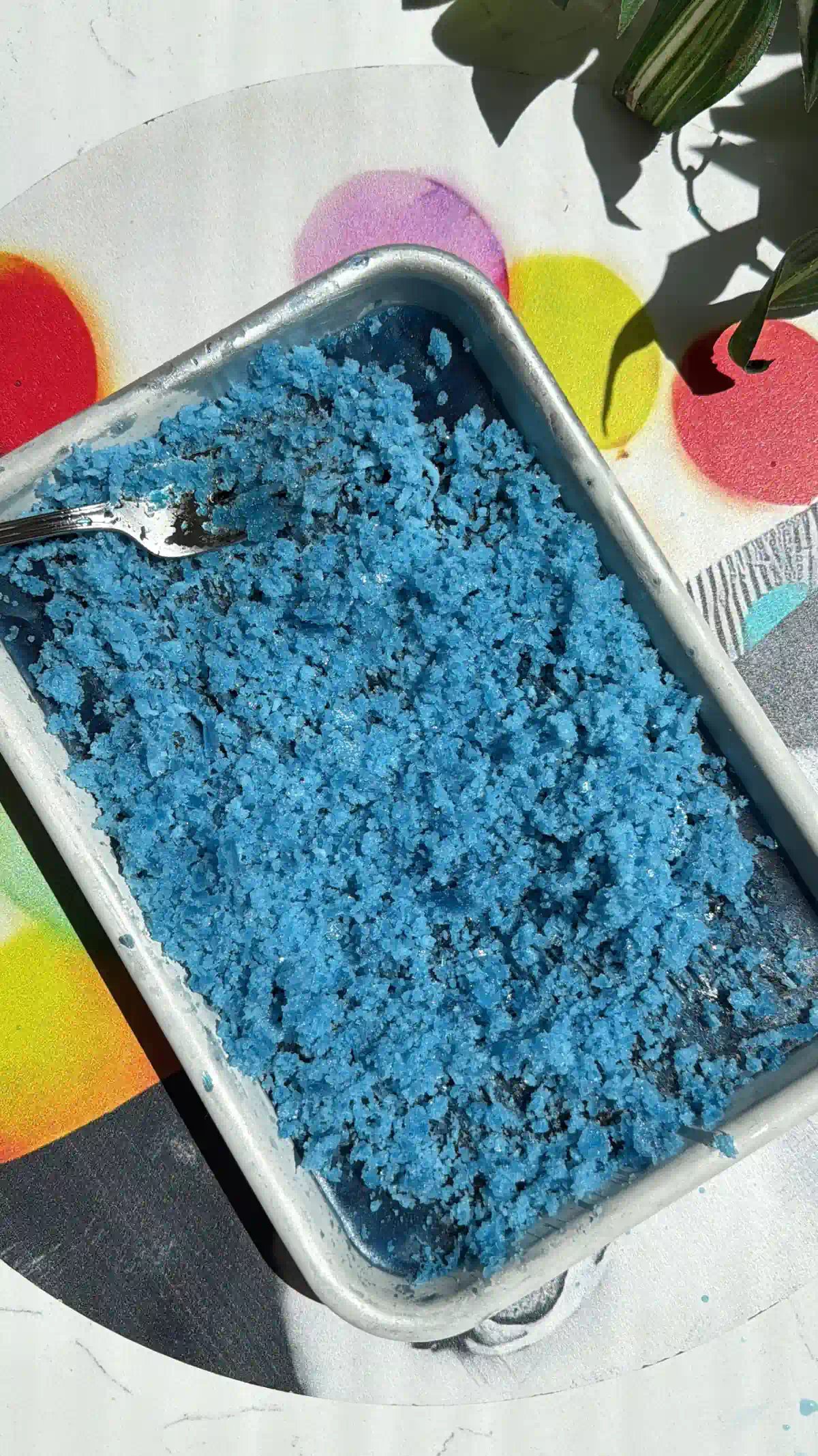 A rectangular metal pan filled with bright blue, scraped butterfly pea granita with a fork resting inside.