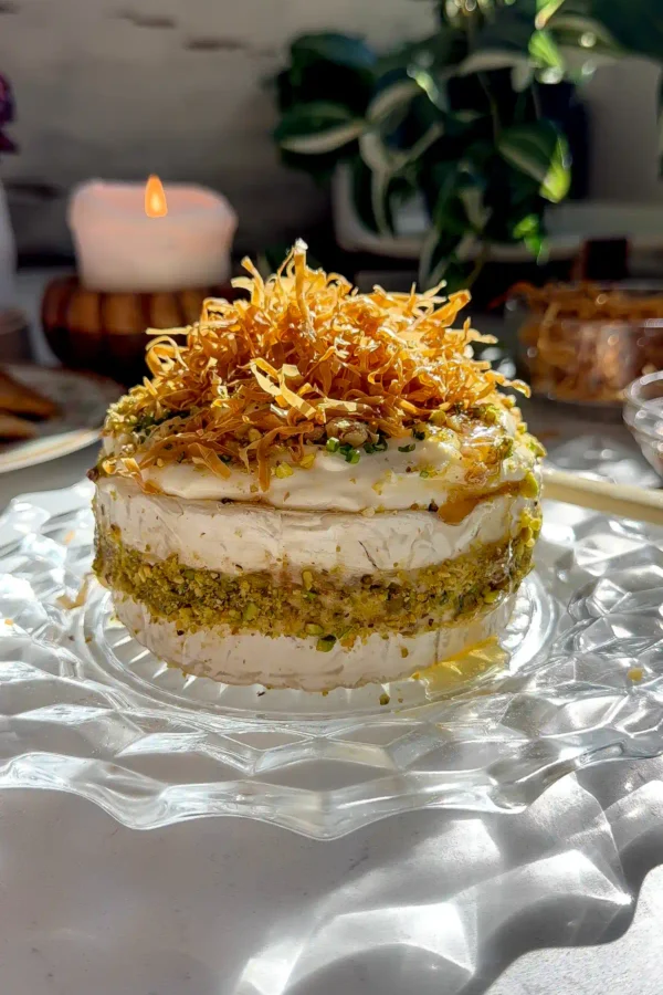 Baklava Brie Cake topped with golden crispy phyllo strands, crushed pistachios, whipped brie, and a honey drizzle on a glass cake stand
