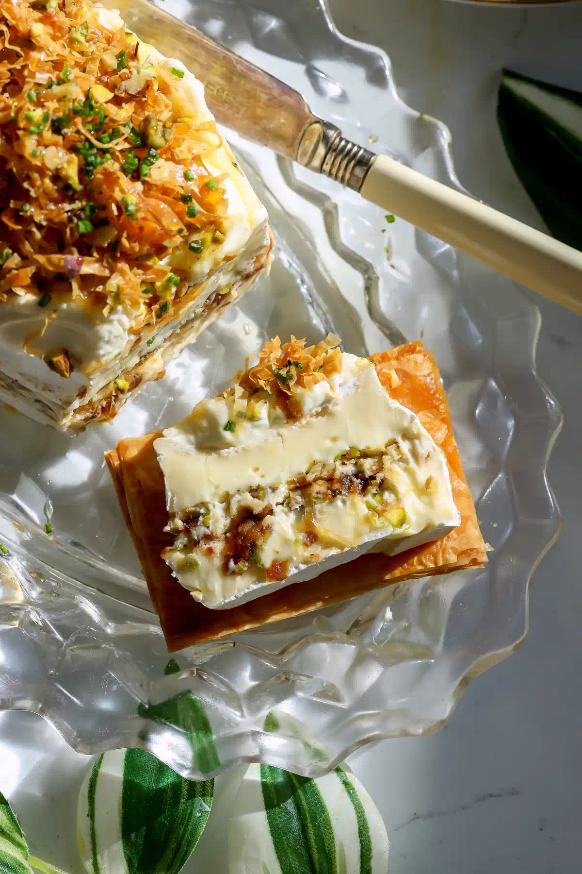 Slice of Baklava Brie Cake on a honey butter phyllo crisp, showing the whipped brie and pistachio walnut date filling, topped with crispy phyllo strands and chives