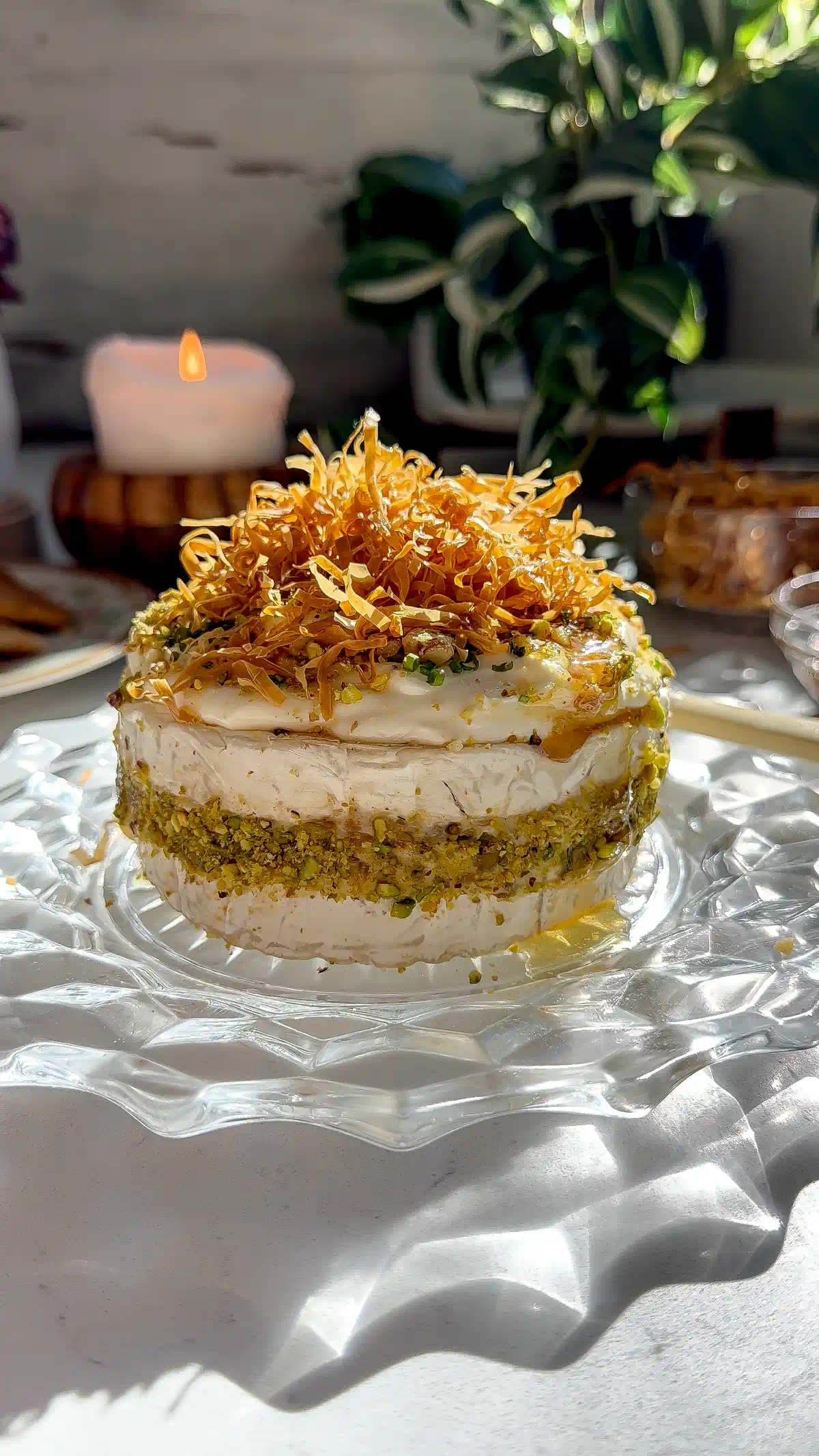 Baklava Brie Cake topped with golden crispy phyllo strands, crushed pistachios, whipped brie, and a honey drizzle on a glass cake stand